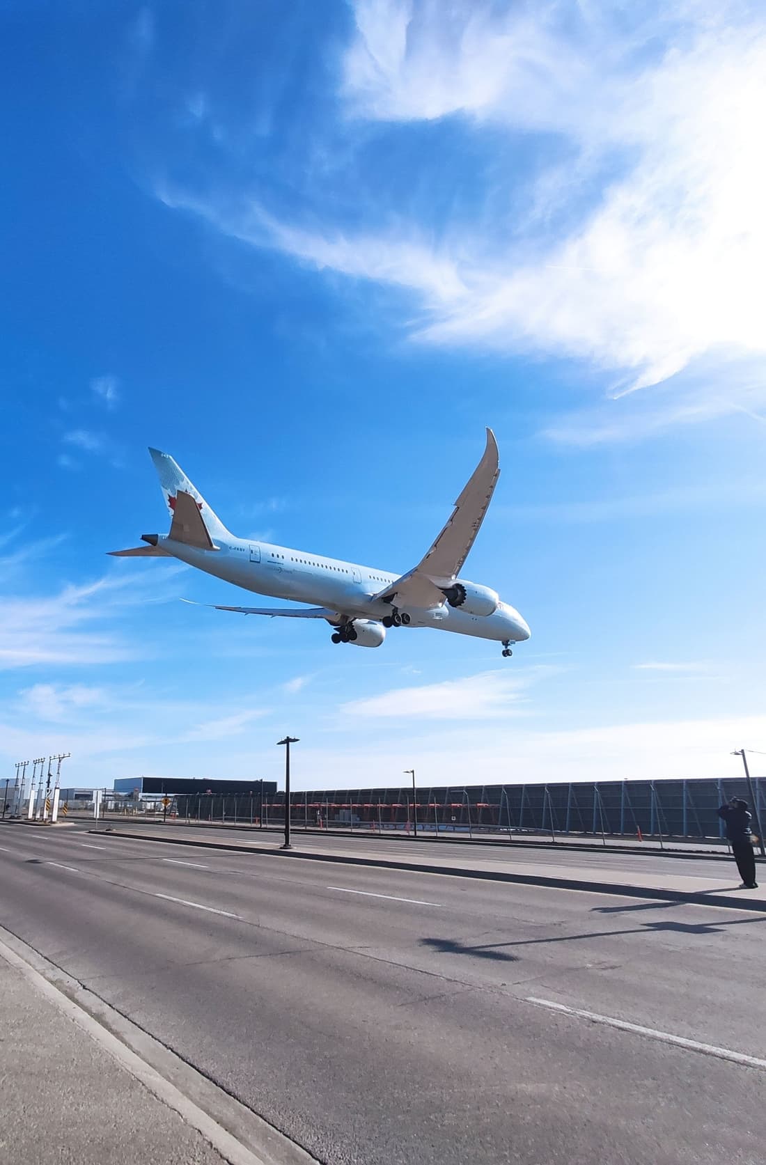 An Air Canada Boeing 787-9 seen from below, gear down and wings flexed upward, low over an empty road beside the airfield fence under a bright blue sky.