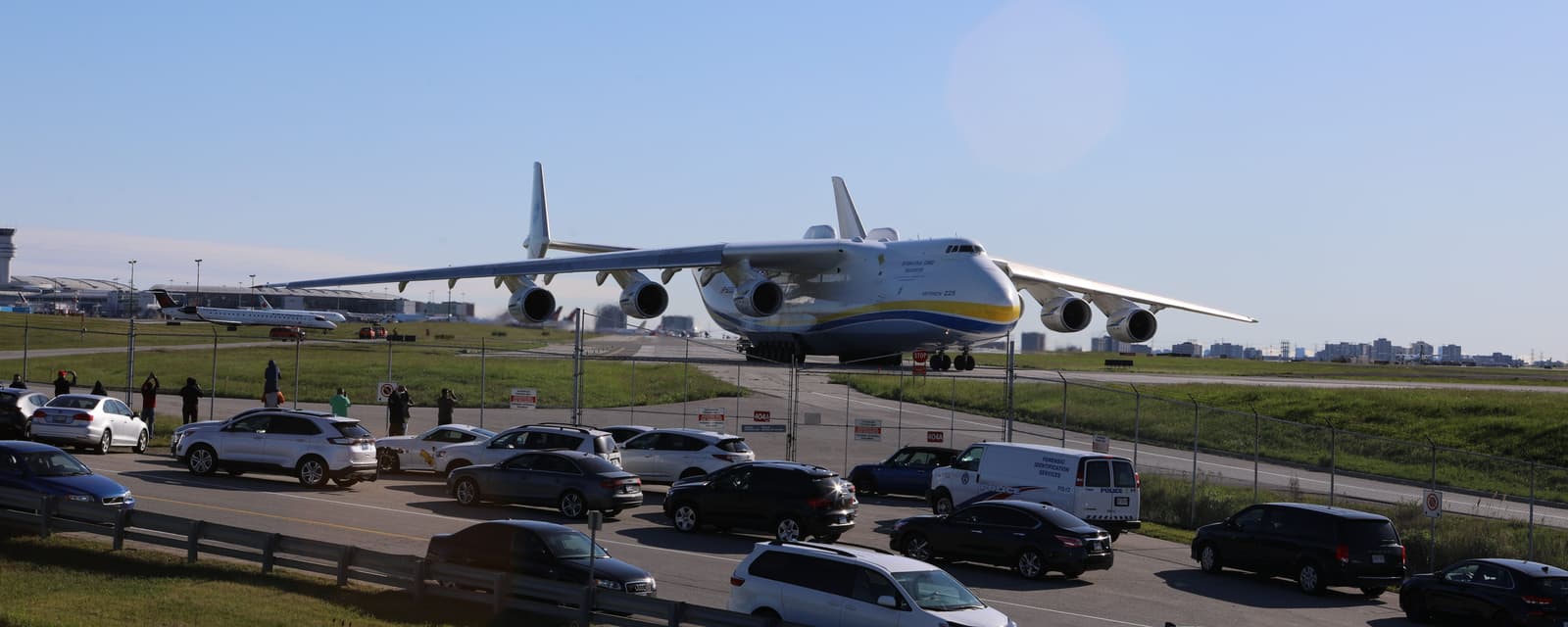 The six-engined Antonov An-225 Mriya on a Pearson taxiway, white with a blue and yellow cheatline, as rows of parked cars and onlookers line the fence in the foreground.