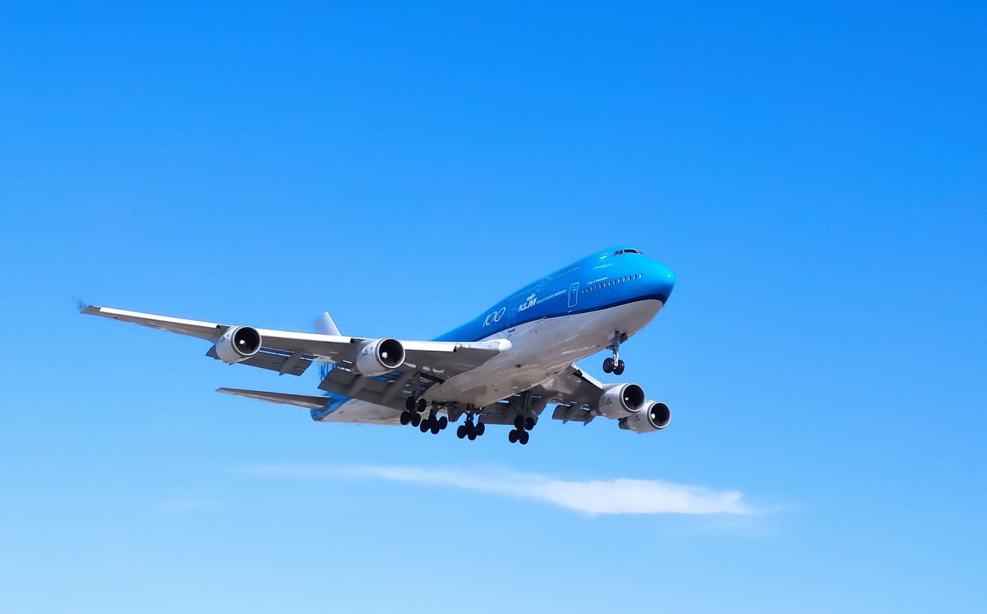 A KLM Boeing 747-400 in light blue, with the airline’s centenary “100” titles, on approach with its landing gear down against a clear sky.