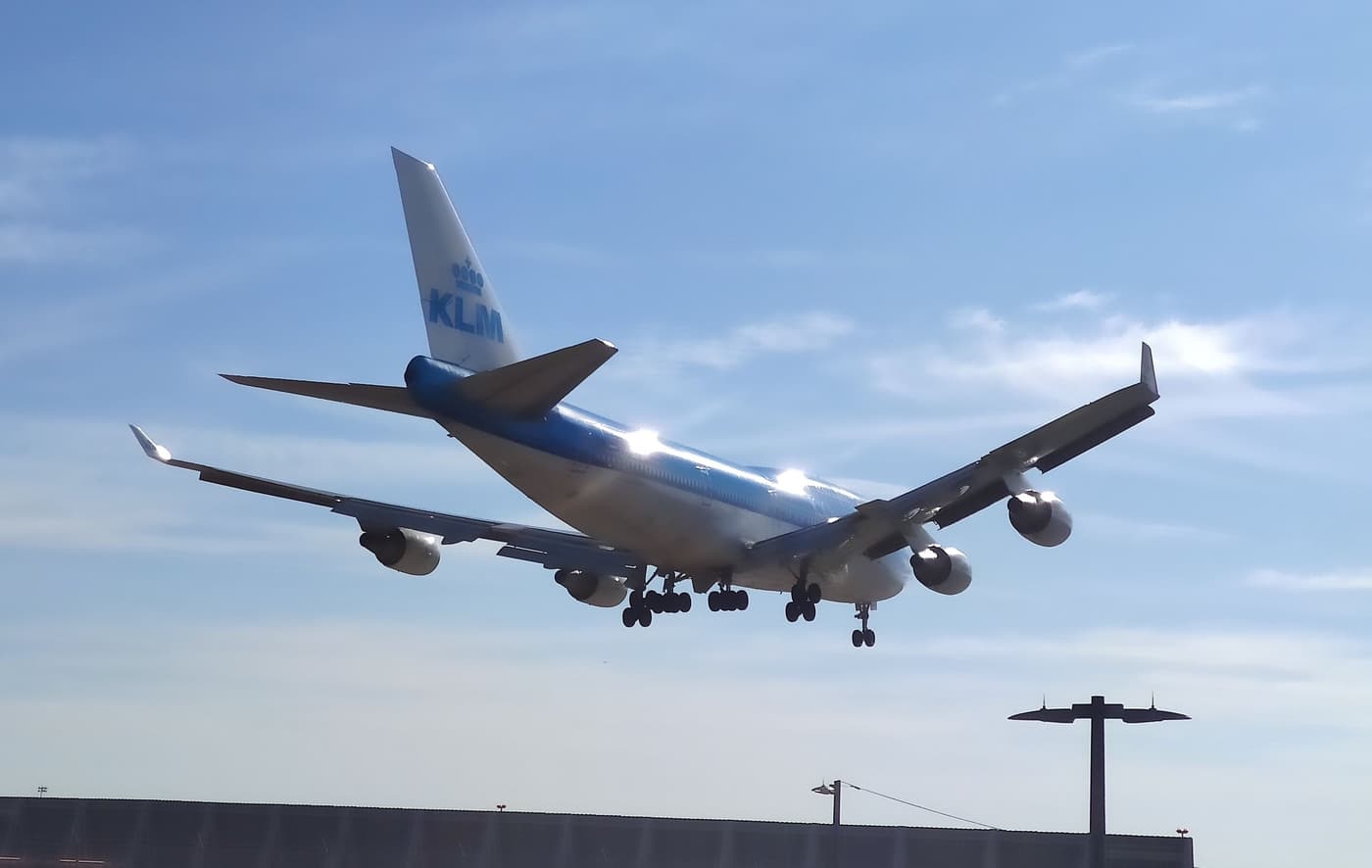 The same KLM 747-400 from behind and below as it passes overhead, sunlight glinting off the fuselage, a light pole in the corner of the frame.
